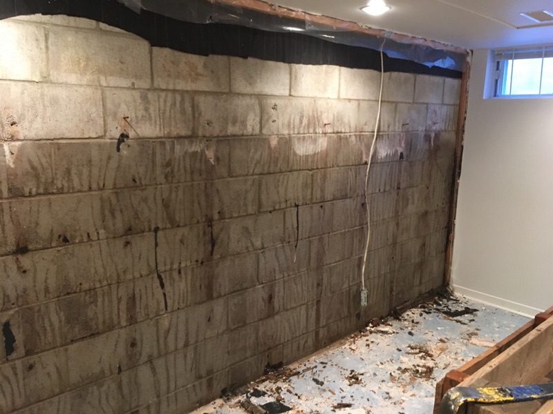 A partially demolished wall in a basement shows exposed concrete blocks and scattered debris on the floor, reminiscent of an urban renewal project. A window near the ceiling lets in natural light, while an electrical cable from City Wide Group hangs loosely from the wall. Waterproofing Company | Basement Leak Repair | City Wide Group | Professional waterproofing company and basement leak repair in Toronto since 1961. | https://www.citywidegroup.com | +1 416-283-5500