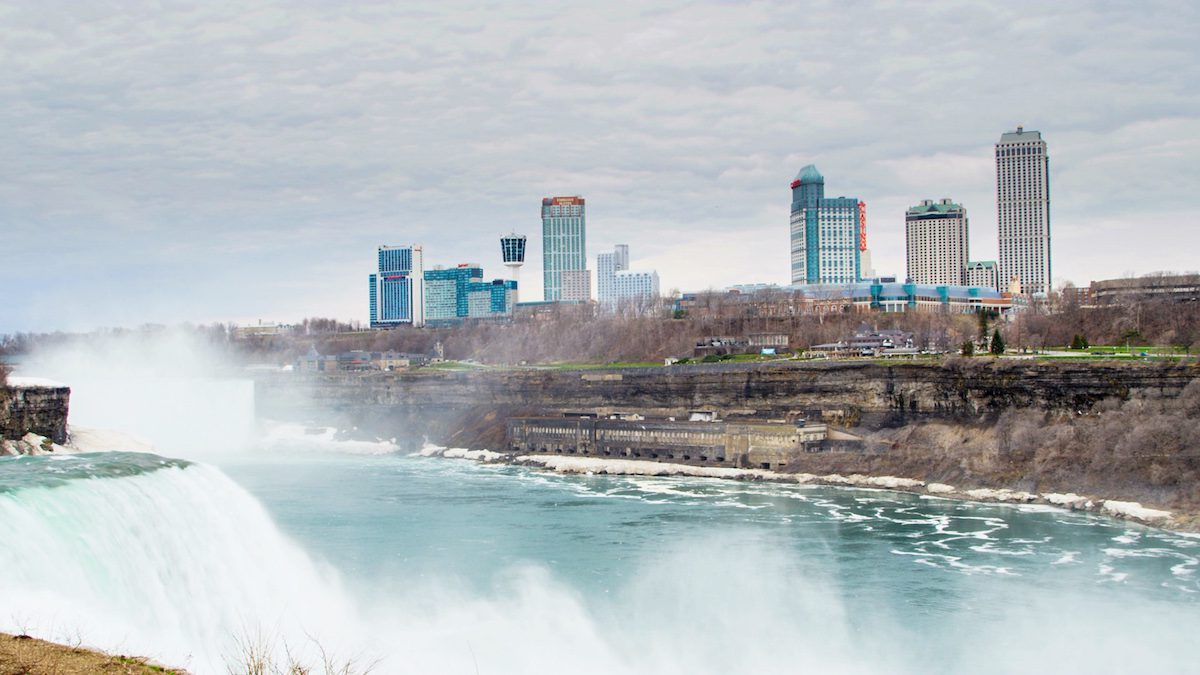 Niagara Falls with mist rising from the waterfall on the left. The skyline of a city with several tall buildings is visible in the background under a cloudy sky. The river flows toward the foreground. Waterproofing Company | Basement Leak Repair | City Wide Group | Professional waterproofing company and basement leak repair in Toronto since 1961. | https://www.citywidegroup.com | +1 416-283-5500