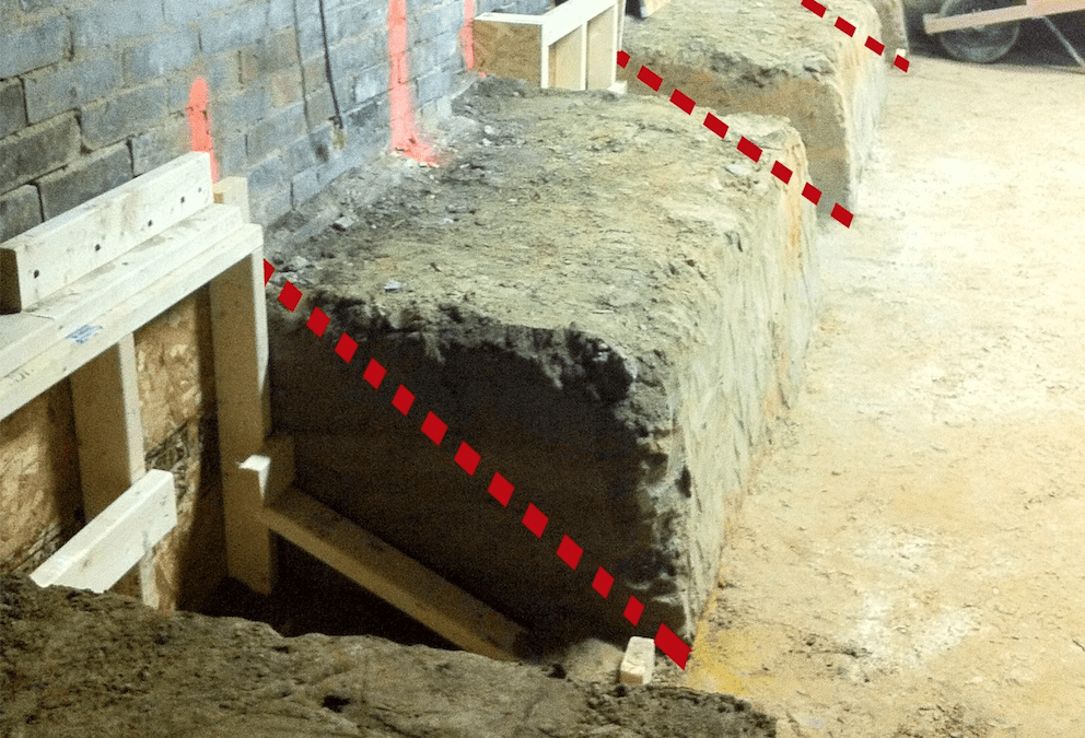 On a construction site, an excavated trench runs alongside a brick wall, supported by wooden braces. Red dashed lines highlight areas of concern or measurement. Dirt and debris scatter the ground—a scene that City Wide Group expertly navigates in their projects across town. Waterproofing Company | Basement Leak Repair | City Wide Group | Professional waterproofing company and basement leak repair in Toronto since 1961. | https://www.citywidegroup.com | +1 416-283-5500
