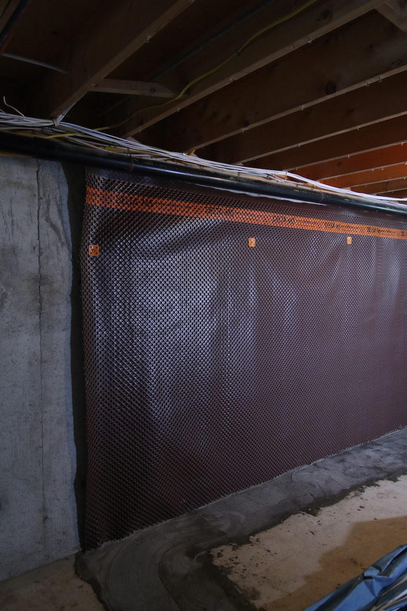 A basement wall with a waterproofing drainage membrane installed. The membrane is black and textured, extending from the floor to the wooden ceiling joists. Wires and a beam are visible at the top. The floor is unfinished concrete. Waterproofing Company | Basement Leak Repair | City Wide Group | Professional waterproofing company and basement leak repair in Toronto since 1961. | https://www.citywidegroup.com | +1 416-283-5500