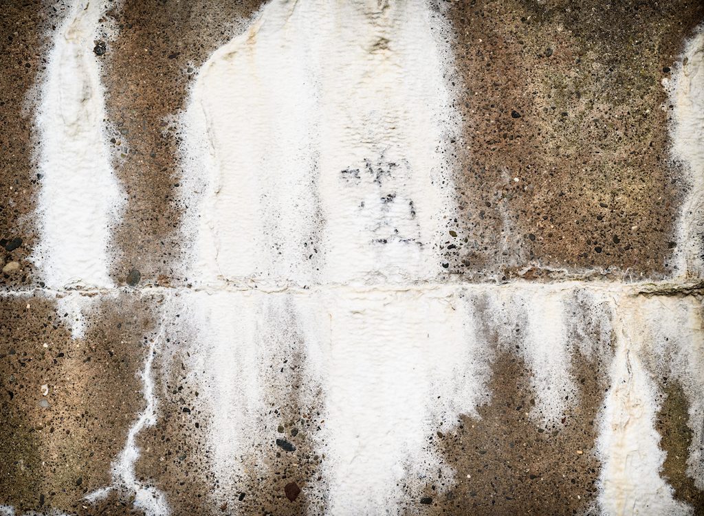 A weathered concrete wall with streaks of white deposits running vertically, resembling water stains or mineral buildup. The surface has a rough texture with visible grain and small pebbles embedded within. Waterproofing Company | Basement Leak Repair | City Wide Group | Professional waterproofing company and basement leak repair in Toronto since 1961. | https://www.citywidegroup.com | +1 416-283-5500