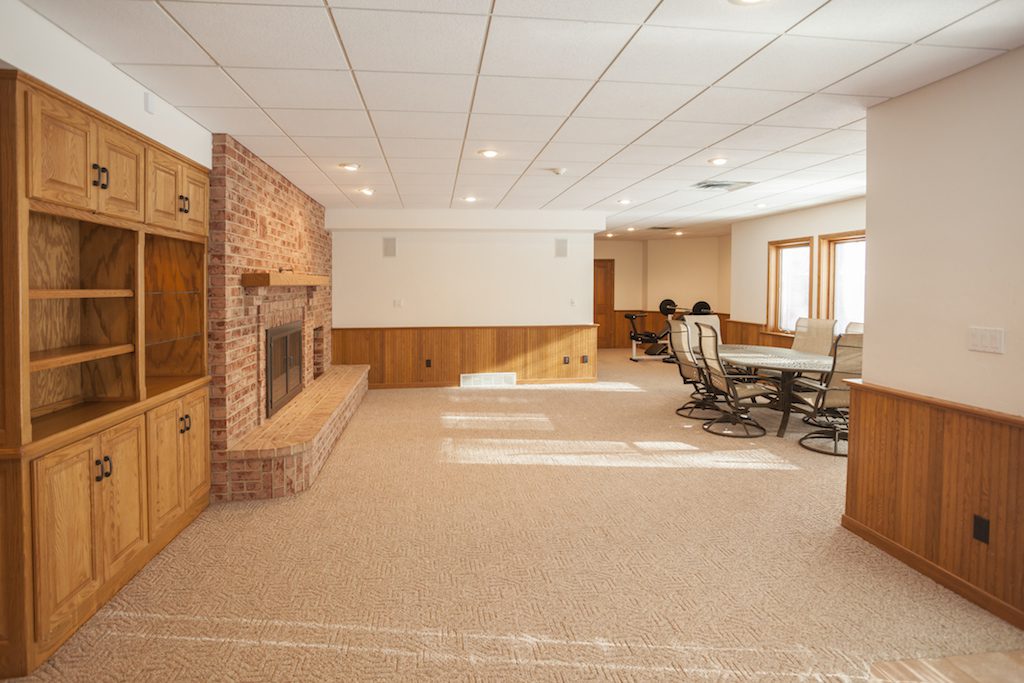 Spacious basement family room with a brick fireplace, wooden cabinetry, carpeted floor, and ceiling lights. Contains a dining table with chairs and exercise equipment near the window. Walls feature wood paneling and there is abundant natural light. Waterproofing Company | Basement Leak Repair | City Wide Group | Professional waterproofing company and basement leak repair in Toronto since 1961. | https://www.citywidegroup.com | +1 416-283-5500