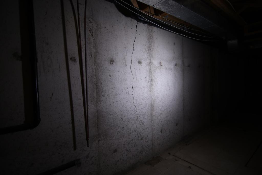 Dimly lit concrete wall in a basement with a visible vertical crack running through the center. Pipes and cables are seen overhead and to the left. The surrounding area is mostly dark. Waterproofing Company | Basement Leak Repair | City Wide Group | Professional waterproofing company and basement leak repair in Toronto since 1961. | https://www.citywidegroup.com | +1 416-283-5500