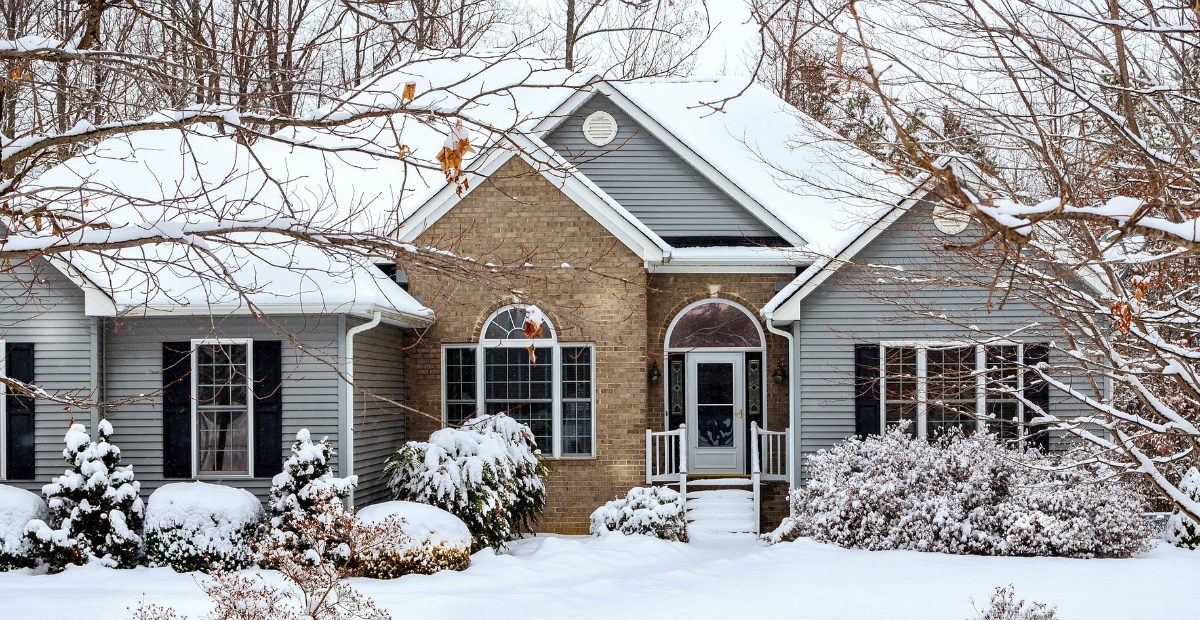 A cozy house with blue siding and a central brick section, surrounded by snow-covered trees and bushes. The front steps and pathway are blanketed in snow, creating a wintery scene. Waterproofing Company | Basement Leak Repair | City Wide Group | Professional waterproofing company and basement leak repair in Toronto since 1961. | https://www.citywidegroup.com | +1 416-283-5500