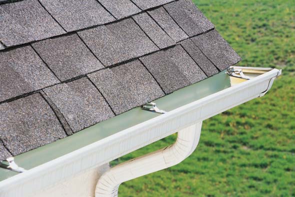 Close-up of a house roof with asphalt shingles and a connected rain gutter. The gutter leads to a white downspout that directs water away from the house. The background shows a grassy area. Waterproofing Company | Basement Leak Repair | City Wide Group | Professional waterproofing company and basement leak repair in Toronto since 1961. | https://www.citywidegroup.com | +1 416-283-5500