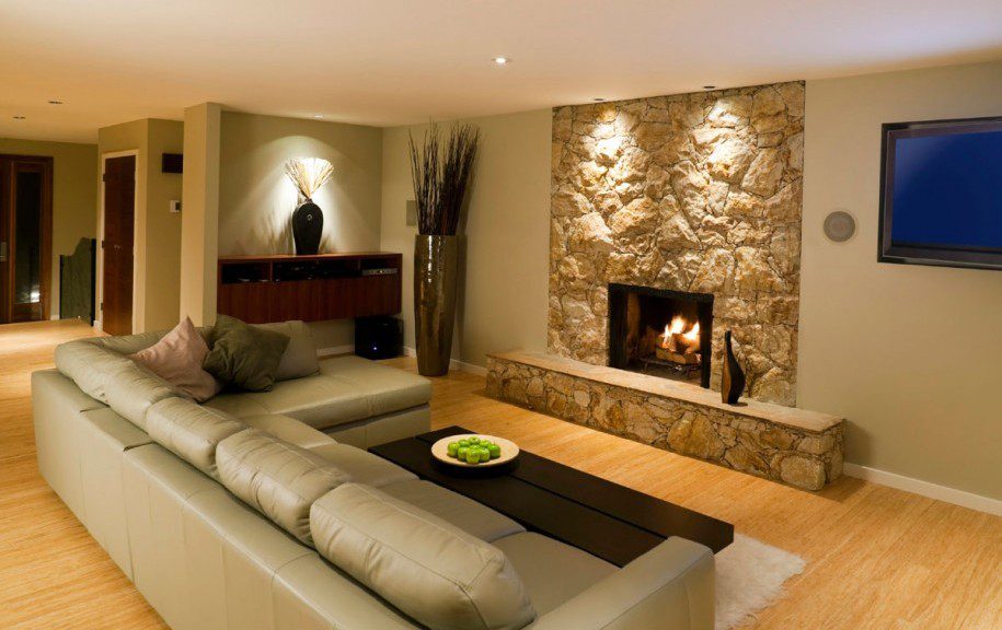 Modern living room with a beige sofa, cushions, and a coffee table holding a bowl of green apples. A stone fireplace is lit, with recessed lights above. A wall-mounted TV is on the right, and decorative tall grass is positioned in a vase. Waterproofing Company | Basement Leak Repair | City Wide Group | Professional waterproofing company and basement leak repair in Toronto since 1961. | https://www.citywidegroup.com | +1 416-283-5500