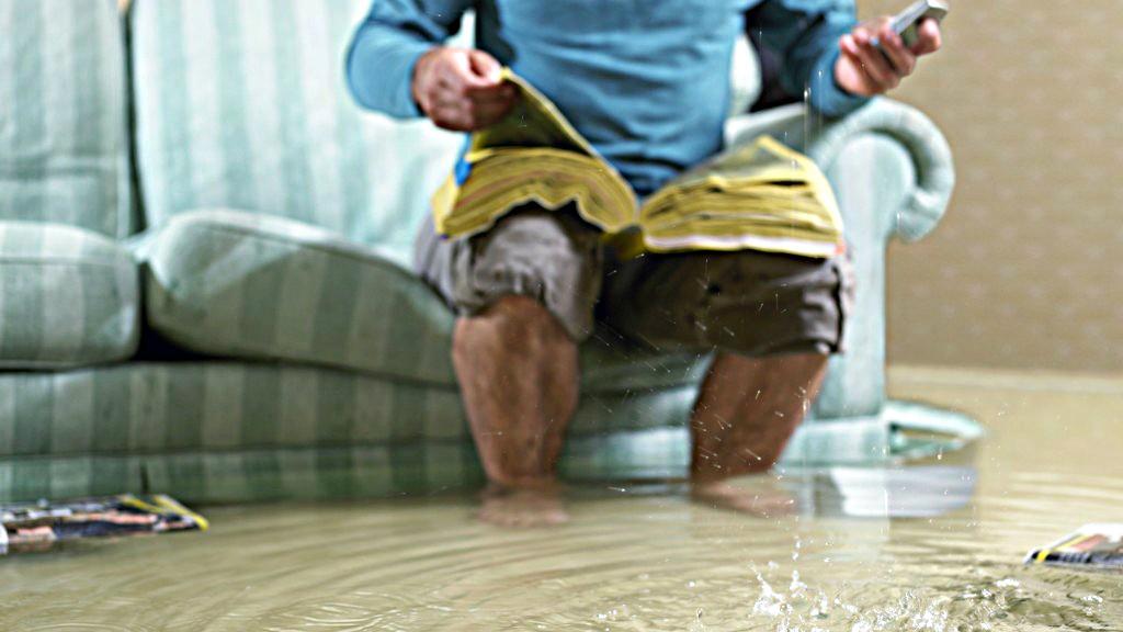 A person sits on a green couch holding a phone and flipping through a yellow book. Water covers the floor, creating ripples. The person wears a blue long-sleeve shirt and brown shorts. A magazine lies on the floor to the left. Waterproofing Company | Basement Leak Repair | City Wide Group | Professional waterproofing company and basement leak repair in Toronto since 1961. | https://www.citywidegroup.com | +1 416-283-5500