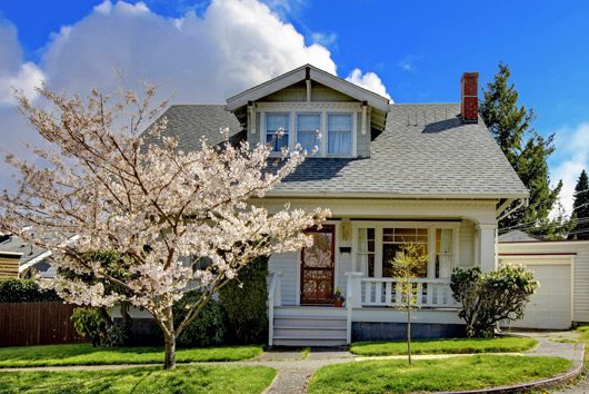 A charming, light gray house with a gabled roof sits under a partly cloudy blue sky. A blooming cherry blossom tree decorates the front yard, and a paved walkway leads to the entrance. A detached garage is visible on the right. Waterproofing Company | Basement Leak Repair | City Wide Group | Professional waterproofing company and basement leak repair in Toronto since 1961. | https://www.citywidegroup.com | +1 416-283-5500