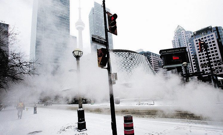 A snowy urban scene with tall buildings partially obscured by steam or fog. The street is empty except for a distant pedestrian. Street signs and a traffic light are visible. A prominent tower rises in the background under a cloudy sky. Waterproofing Company | Basement Leak Repair | City Wide Group | Professional waterproofing company and basement leak repair in Toronto since 1961. | https://www.citywidegroup.com | +1 416-283-5500