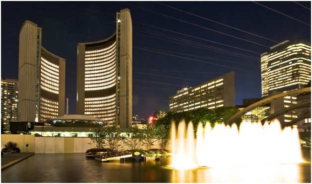 Night view of a modern cityscape with illuminated twin curved buildings, likely a city hall, beside a brightly lit fountain and surrounded by other lit high-rise structures, all reflecting on a calm water surface. Waterproofing Company | Basement Leak Repair | City Wide Group | Professional waterproofing company and basement leak repair in Toronto since 1961. | https://www.citywidegroup.com | +1 416-283-5500