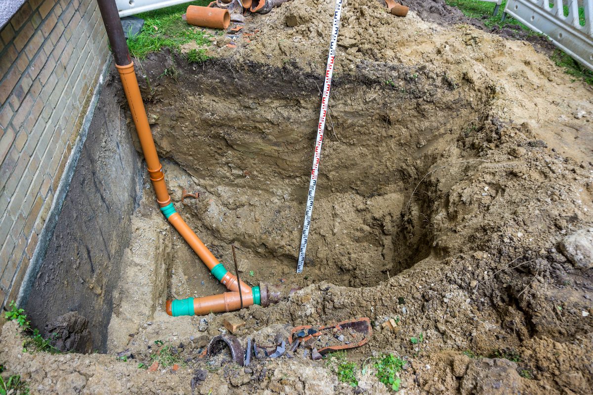 A significant excavation by City Wide Group reveals a network of orange PVC pipes under the building. A measuring rod stands amid broken pieces and scattered dirt, hinting at an ongoing construction effort to improve urban infrastructure. Waterproofing Company | Basement Leak Repair | City Wide Group | Professional waterproofing company and basement leak repair in Toronto since 1961. | https://www.citywidegroup.com | +1 416-283-5500