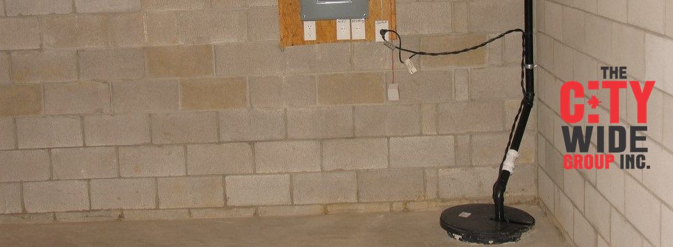 A basement with bare concrete walls and floor. In the corner, a sump pump is installed with pipes running vertically. Electrical outlets and a circuit box are mounted on the wall. The City Wide Group Inc. logo is visible in the lower right corner. Waterproofing Company | Basement Leak Repair | City Wide Group | Professional waterproofing company and basement leak repair in Toronto since 1961. | https://www.citywidegroup.com | +1 416-283-5500
