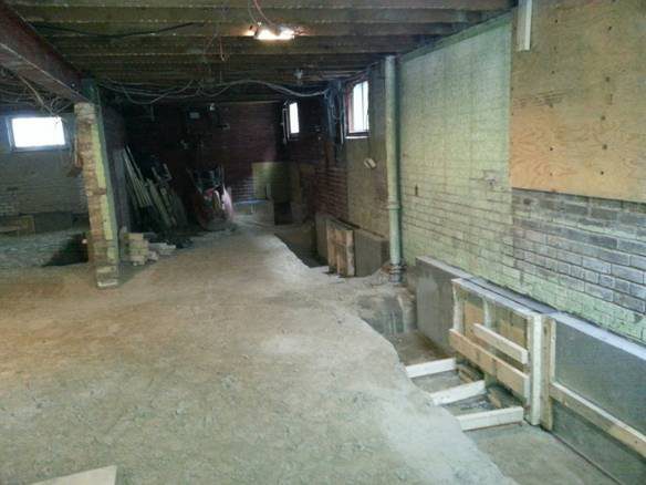 A dimly lit basement under renovation with unfinished floors and exposed ceiling. Building materials like wooden planks and bricks are scattered, with pipes and cables visible on the ceiling and walls. Small windows let in minimal light. Waterproofing Company | Basement Leak Repair | City Wide Group | Professional waterproofing company and basement leak repair in Toronto since 1961. | https://www.citywidegroup.com | +1 416-283-5500