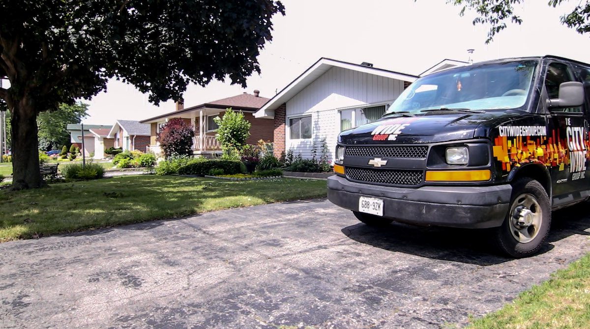 A black Chevrolet van with orange and yellow text from City Wide Group is parked on a driveway in front of a suburban house. The house features a lawn and porch, while trees and shrubs line the street, capturing the essence of a vibrant news day under clear skies. Waterproofing Company | Basement Leak Repair | City Wide Group | Professional waterproofing company and basement leak repair in Toronto since 1961. | https://www.citywidegroup.com | +1 416-283-5500