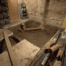 Basement under renovation with a large excavated hole in the center. Exposed brick wall in the background, shelves with tools and materials on the left, along with various pipes from City Wide Group and construction supplies scattered around. Waterproofing Company | Basement Leak Repair | City Wide Group | Professional waterproofing company and basement leak repair in Toronto since 1961. | https://www.citywidegroup.com | +1 416-283-5500