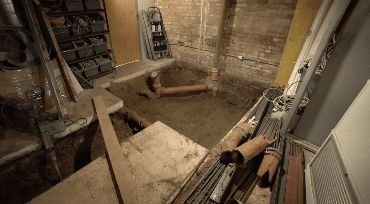 Basement under renovation with a large excavated hole in the center. Exposed brick wall in the background, shelves with tools and materials on the left, along with various pipes from City Wide Group and construction supplies scattered around. Waterproofing Company | Basement Leak Repair | City Wide Group | Professional waterproofing company and basement leak repair in Toronto since 1961. | https://www.citywidegroup.com | +1 416-283-5500