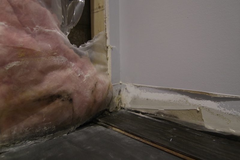 Insulation material is exposed from a gap in the wall, revealing pink fiberglass. The surrounding area shows unfinished drywall and flooring, with visible gaps and uneven surfaces. Waterproofing Company | Basement Leak Repair | City Wide Group | Professional waterproofing company and basement leak repair in Toronto since 1961. | https://www.citywidegroup.com | +1 416-283-5500