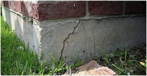 A close-up of a buildings foundation corner showing visible cracks and wear in the concrete. There are red bricks on top of the foundation. Grass and a fallen leaf are visible at the base. Waterproofing Company | Basement Leak Repair | City Wide Group | Professional waterproofing company and basement leak repair in Toronto since 1961. | https://www.citywidegroup.com | +1 416-283-5500