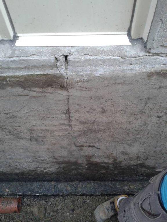 Cracked concrete wall beneath a window, with visible damage extending from the window frame downward. A person wearing work boots and gray pants stands nearby on a gravel surface. Waterproofing Company | Basement Leak Repair | City Wide Group | Professional waterproofing company and basement leak repair in Toronto since 1961. | https://www.citywidegroup.com | +1 416-283-5500