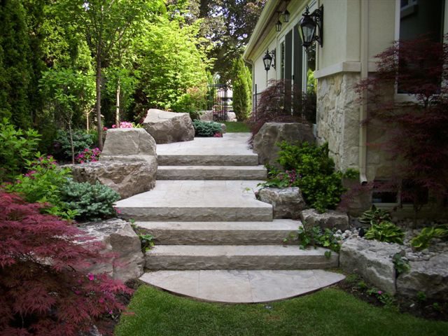 Stone steps lead up through a lush garden with various shrubs, trees, and flowers on either side. Large rocks flank the pathway, and a building with lanterns is on the right. The area appears well-maintained and tranquil. Waterproofing Company | Basement Leak Repair | City Wide Group | Professional waterproofing company and basement leak repair in Toronto since 1961. | https://www.citywidegroup.com | +1 416-283-5500
