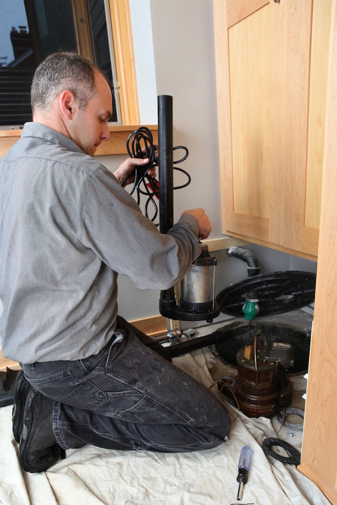 A person kneels on a cloth, expertly repairing plumbing under a sink for City Wide Group. Tools in hand, they focus on a cylindrical component, with wooden cabinetry open above them. Waterproofing Company | Basement Leak Repair | City Wide Group | Professional waterproofing company and basement leak repair in Toronto since 1961. | https://www.citywidegroup.com | +1 416-283-5500