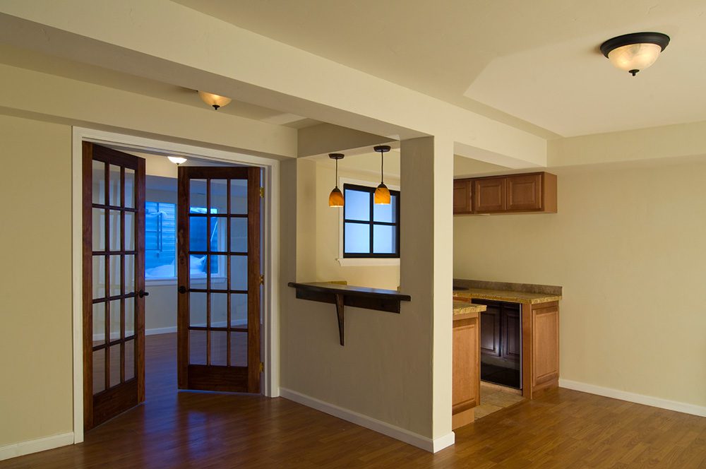 The image shows an empty room with hardwood floors and cream walls. There are two doorways with glass-paneled doors, a small bar area with a countertop and pendant lights, and a window above a set of wooden cabinets. The room is well-lit. Waterproofing Company | Basement Leak Repair | City Wide Group | Professional waterproofing company and basement leak repair in Toronto since 1961. | https://www.citywidegroup.com | +1 416-283-5500