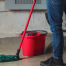 Person wearing jeans and black shoes standing on a wet concrete floor with a red mop bucket and a green mop. Pipes and a gray wall are in the background. Waterproofing Company | Basement Leak Repair | City Wide Group | Professional waterproofing company and basement leak repair in Toronto since 1961. | https://www.citywidegroup.com | +1 416-283-5500