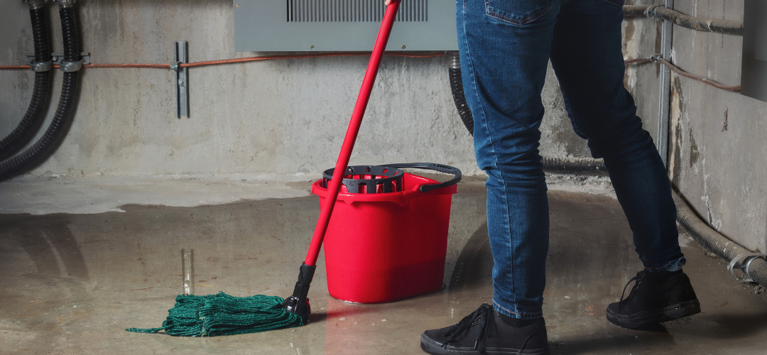 Person wearing jeans and black shoes standing on a wet concrete floor with a red mop bucket and a green mop. Pipes and a gray wall are in the background. Waterproofing Company | Basement Leak Repair | City Wide Group | Professional waterproofing company and basement leak repair in Toronto since 1961. | https://www.citywidegroup.com | +1 416-283-5500