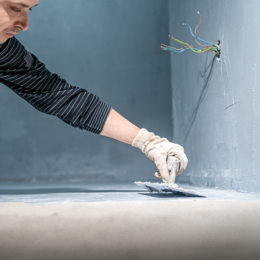 A person from the City Wide Group is applying plaster to a wall with a trowel, focused on smoothing the surface while wearing gloves and a striped shirt. Exposed wires are visible near the top right corner, suggesting this construction site involves basement waterproofing. Waterproofing Company | Basement Leak Repair | City Wide Group | Professional waterproofing company and basement leak repair in Toronto since 1961. | https://www.citywidegroup.com | +1 416-283-5500