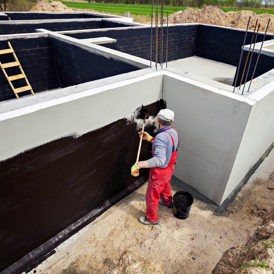 A construction worker in red overalls from City Wide Group expertly applies waterproofing tar to the foundation walls. The partially painted walls hint at progress, while a bucket of tar and a ladder stand ready, showcasing the professional touch of this basement waterproofing company. Waterproofing Company | Basement Leak Repair | City Wide Group | Professional waterproofing company and basement leak repair in Toronto since 1961. | https://www.citywidegroup.com | +1 416-283-5500