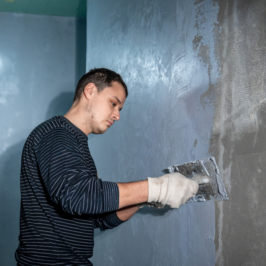 In a pristine basement, a person donning a striped shirt and gloves expertly uses a trowel to apply plaster on a blue wall, channeling the precision expected from City Wide Group, renowned for their excellence in basement waterproofing. Waterproofing Company | Basement Leak Repair | City Wide Group | Professional waterproofing company and basement leak repair in Toronto since 1961. | https://www.citywidegroup.com | +1 416-283-5500