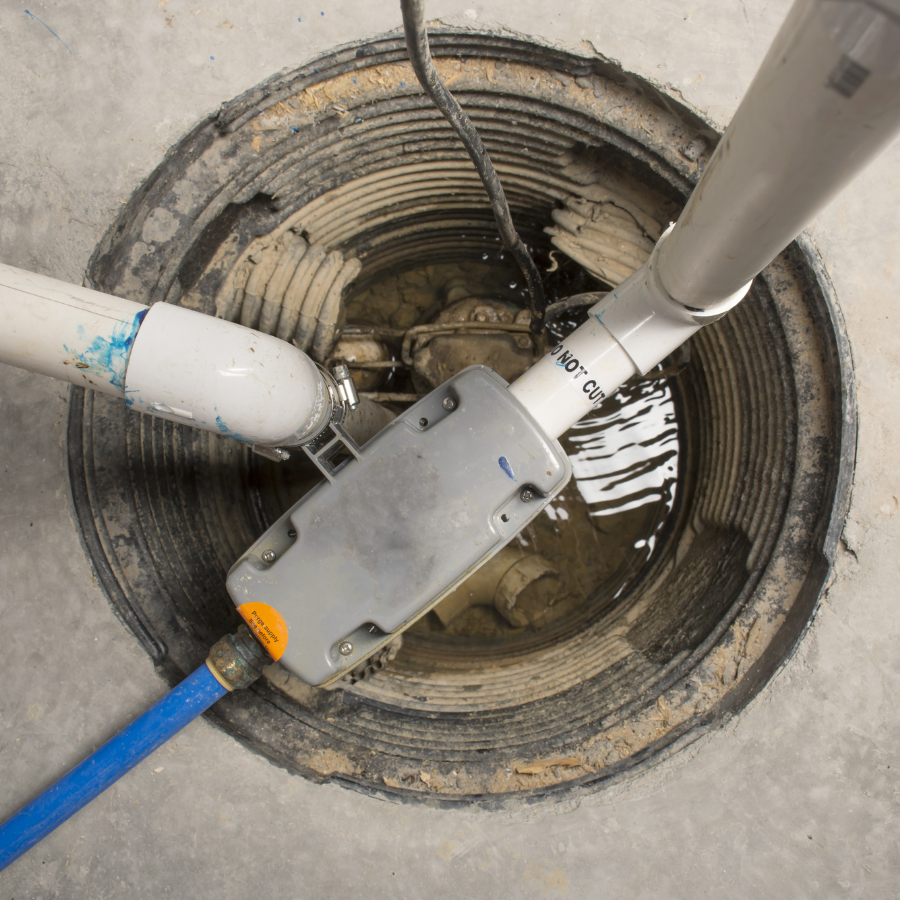Close-up of a sump pump pit with PVC pipes and water inside, expertly installed by City Wide Group, a premier basement waterproofing company. The white pipes connect to a gray control box, nestled into the concrete floor, suggesting a basement or utility area setup. Waterproofing Company | Basement Leak Repair | City Wide Group | Professional waterproofing company and basement leak repair in Toronto since 1961. | https://www.citywidegroup.com | +1 416-283-5500