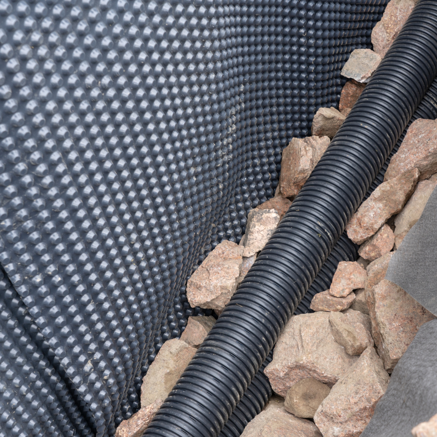 Close-up of a perforated drainage pipe surrounded by rocks and gravel. The background reveals a dimpled waterproofing membrane on the wall, expertly installed by Basement Waterproofing Company City Wide Group. Waterproofing Company | Basement Leak Repair | City Wide Group | Professional waterproofing company and basement leak repair in Toronto since 1961. | https://www.citywidegroup.com | +1 416-283-5500
