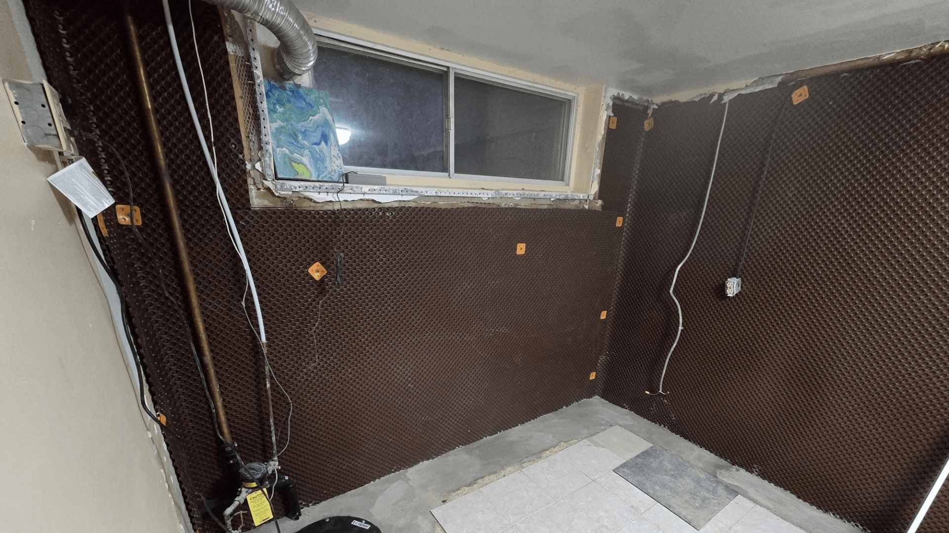 Partially finished basement room with brown dimpled waterproofing membrane on the walls, exposed pipes, electrical wires, and an uncovered window. The floor is unfinished with some tiles placed in one corner. Waterproofing Company | Basement Leak Repair | City Wide Group | Professional waterproofing company and basement leak repair in Toronto since 1961. | https://www.citywidegroup.com | +1 416-283-5500