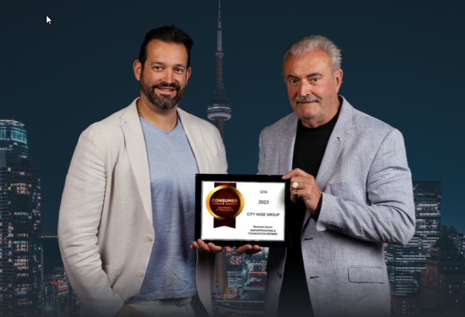 Two men in suits hold an award plaque together in front of a dark, cityscape background with a prominent lit-up tower. The plaque is from the Consumer Choice Award 2023. Waterproofing Company | Basement Leak Repair | City Wide Group | Professional waterproofing company and basement leak repair in Toronto since 1961. | https://www.citywidegroup.com | +1 416-283-5500