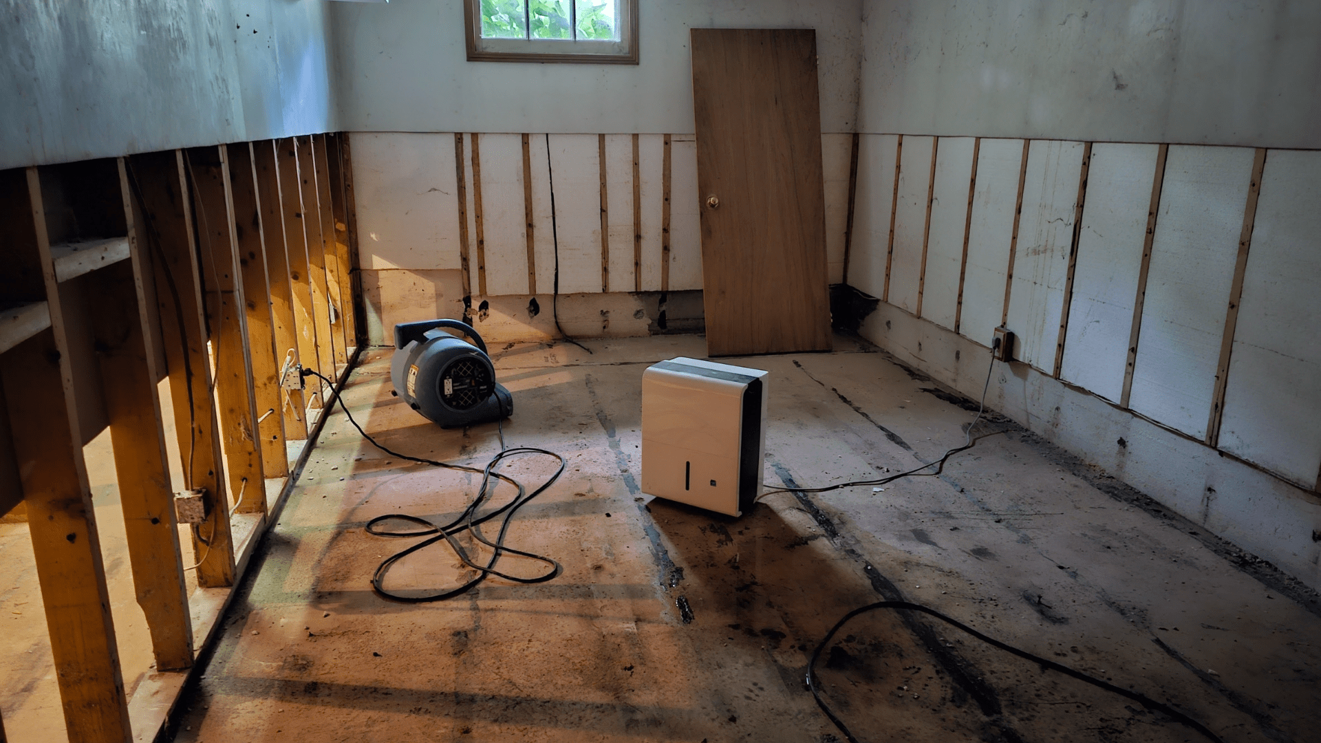 A bare, partially stripped room with exposed wall studs and no flooring. A fan and a dehumidifier are on the floor, with cords trailing. A small window lets in light and a detached door leans against the far wall. Waterproofing Company | Basement Leak Repair | City Wide Group | Professional waterproofing company and basement leak repair in Toronto since 1961. | https://www.citywidegroup.com | +1 416-283-5500