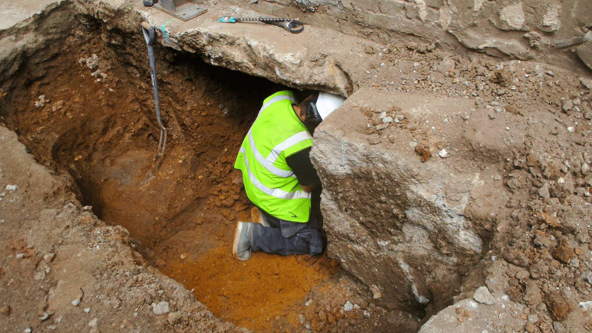 A construction worker in a high-visibility vest and hard hat kneels in a deep, dug-out trench, partially covered by a slab of concrete, working or inspecting something underground. Waterproofing Company | Basement Leak Repair | City Wide Group | Professional waterproofing company and basement leak repair in Toronto since 1961. | https://www.citywidegroup.com | +1 416-283-5500
