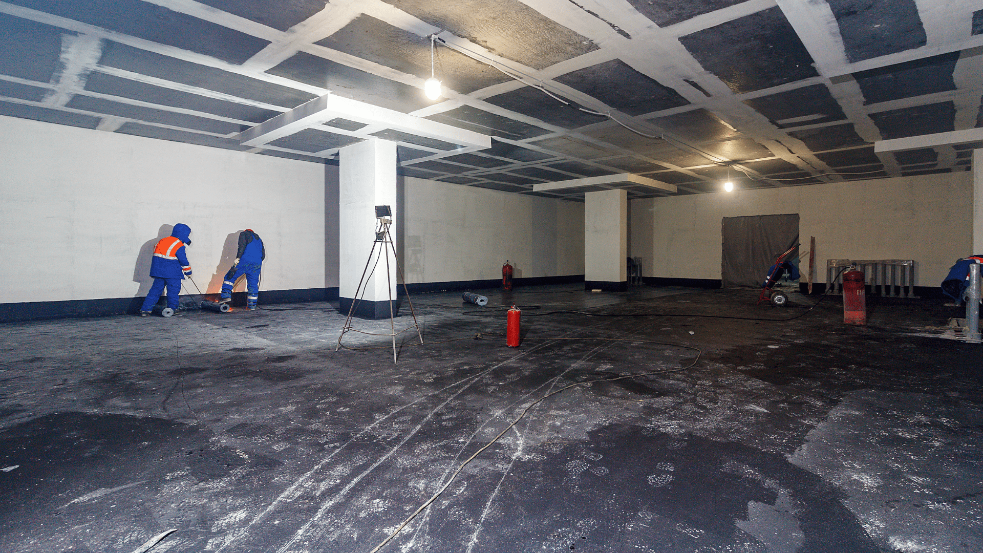 Several workers in safety gear are cleaning or working on the floor of a large, unfinished indoor space with exposed ceiling tiles and columns. Construction materials and fire extinguishers are visible around the room. Waterproofing Company | Basement Leak Repair | City Wide Group | Professional waterproofing company and basement leak repair in Toronto since 1961. | https://www.citywidegroup.com | +1 416-283-5500