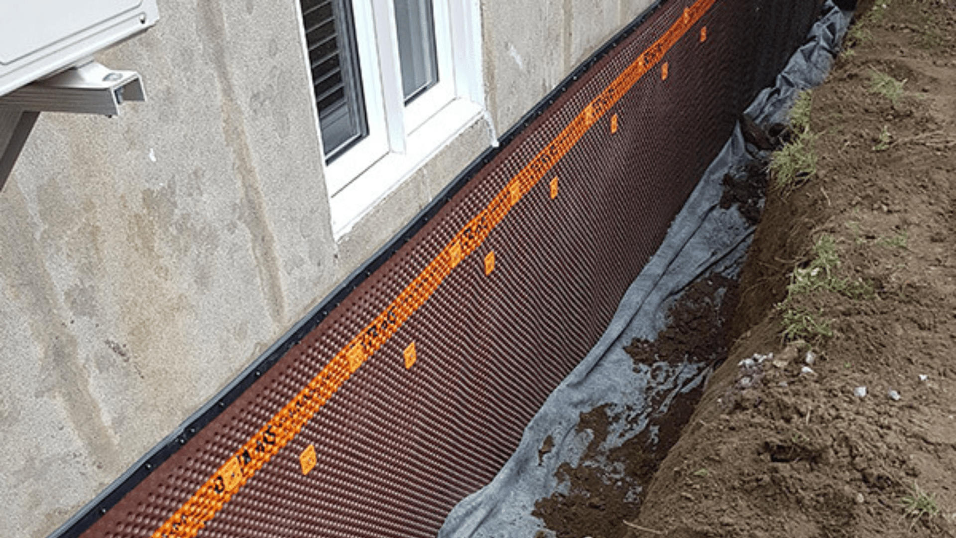 A foundation wall is covered with a dimpled waterproof membrane held in place by orange fasteners. The ground beside the wall has been excavated, exposing soil and part of a house window above. Waterproofing Company | Basement Leak Repair | City Wide Group | Professional waterproofing company and basement leak repair in Toronto since 1961. | https://www.citywidegroup.com | +1 416-283-5500