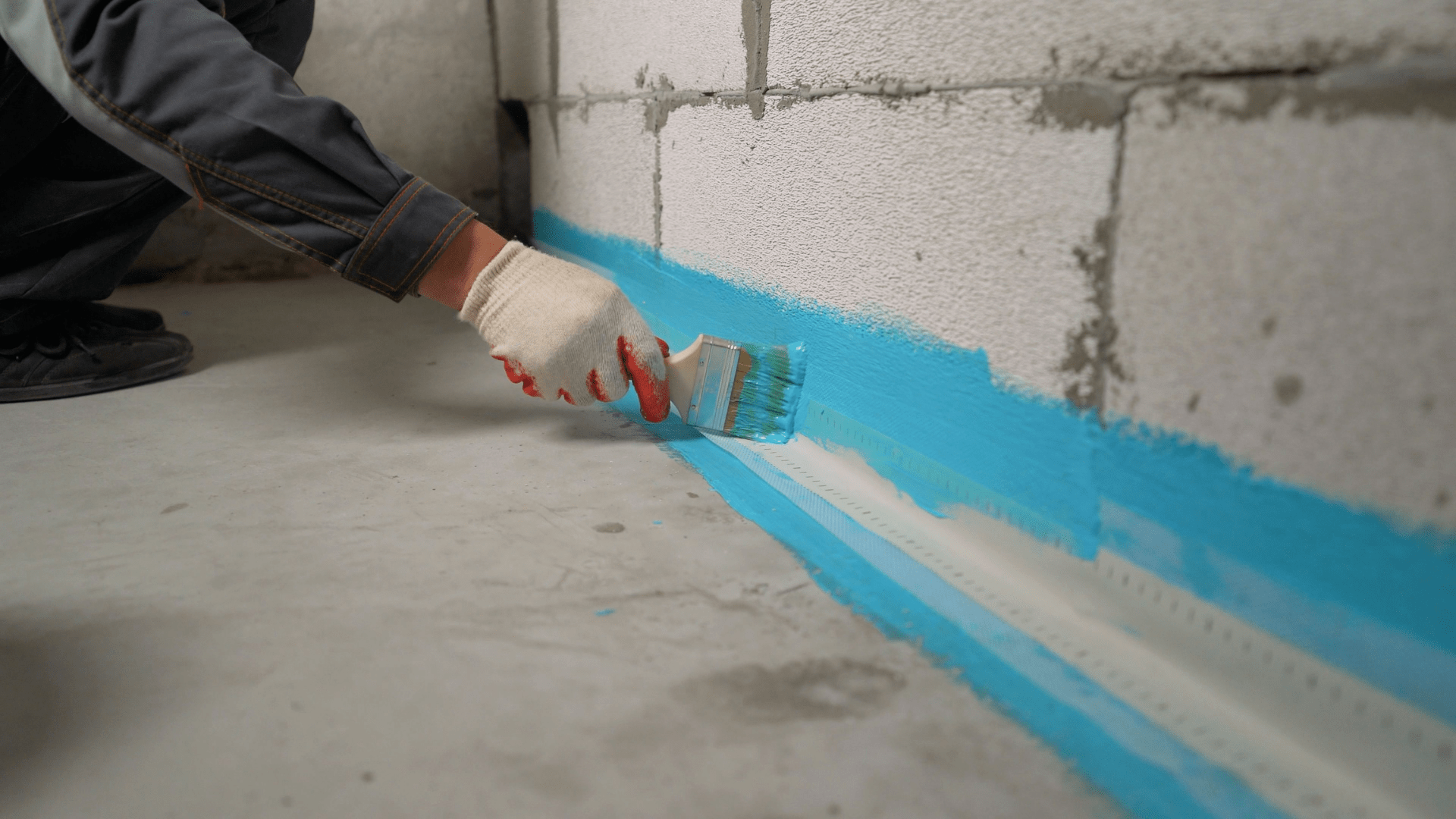 A person wearing a glove uses a paintbrush to apply a blue waterproofing sealant along the base of a concrete wall and floor in a building under construction. Waterproofing Company | Basement Leak Repair | City Wide Group | Professional waterproofing company and basement leak repair in Toronto since 1961. | https://www.citywidegroup.com | +1 416-283-5500