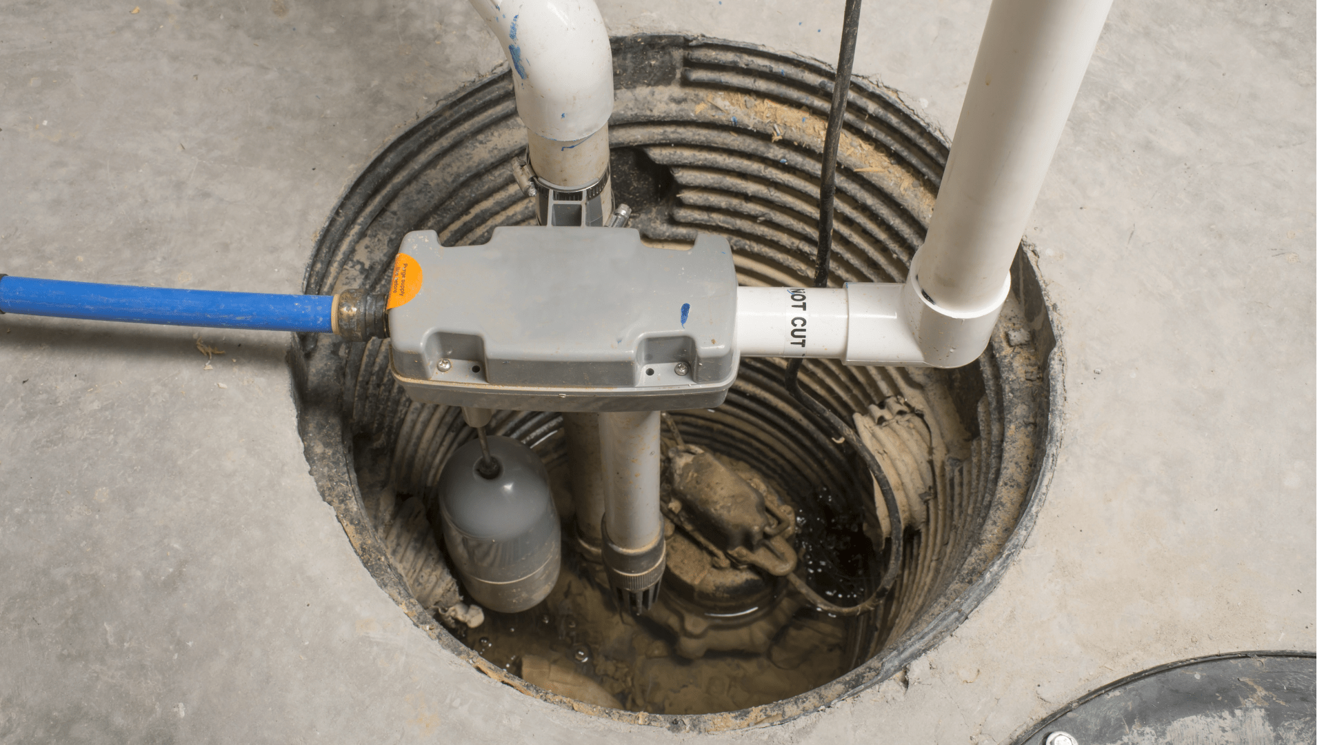A sump pump system installed in a basement floor pit, with connected white PVC pipes, a blue hose, and a float switch mechanism visible inside the circular pit. Waterproofing Company | Basement Leak Repair | City Wide Group | Professional waterproofing company and basement leak repair in Toronto since 1961. | https://www.citywidegroup.com | +1 416-283-5500