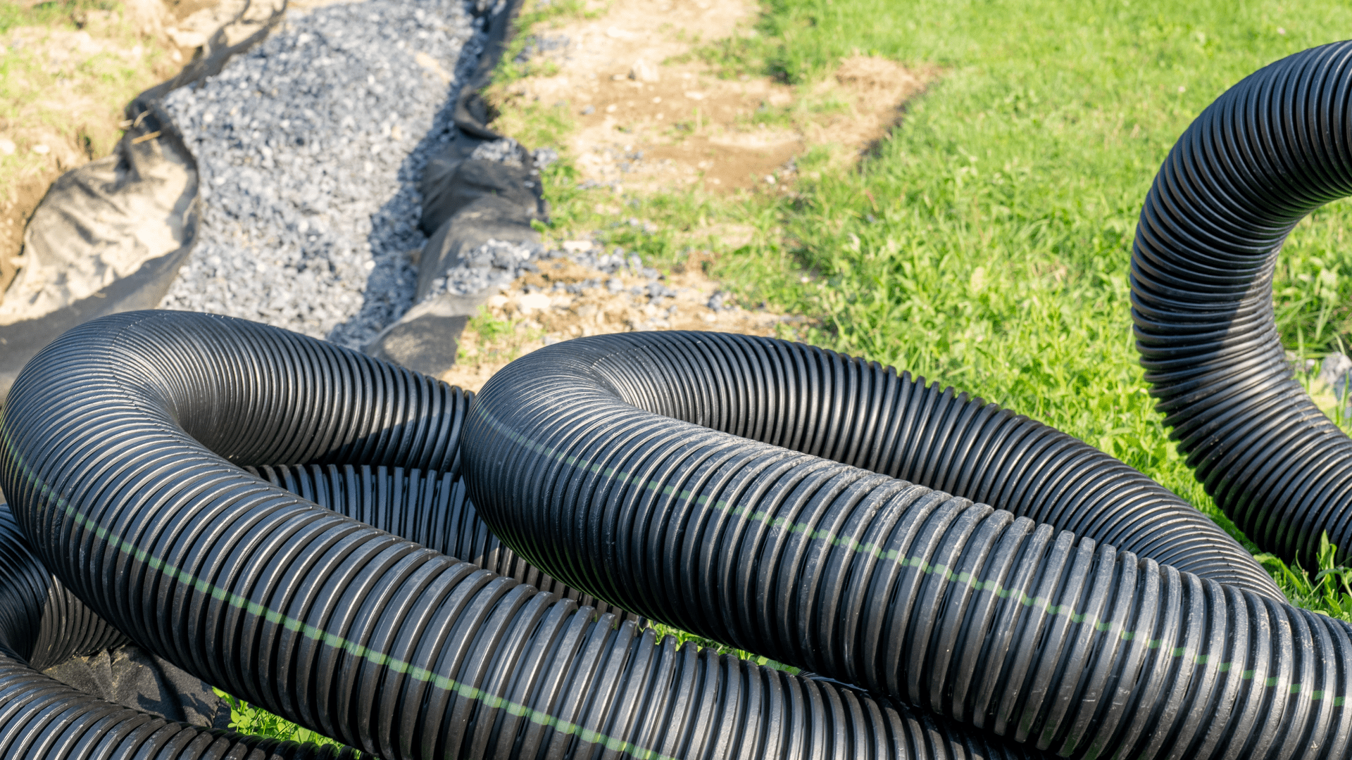 Large black corrugated drainage pipes are coiled on green grass near a trench filled with gravel, likely prepared for installation as part of a landscape drainage system. Waterproofing Company | Basement Leak Repair | City Wide Group | Professional waterproofing company and basement leak repair in Toronto since 1961. | https://www.citywidegroup.com | +1 416-283-5500