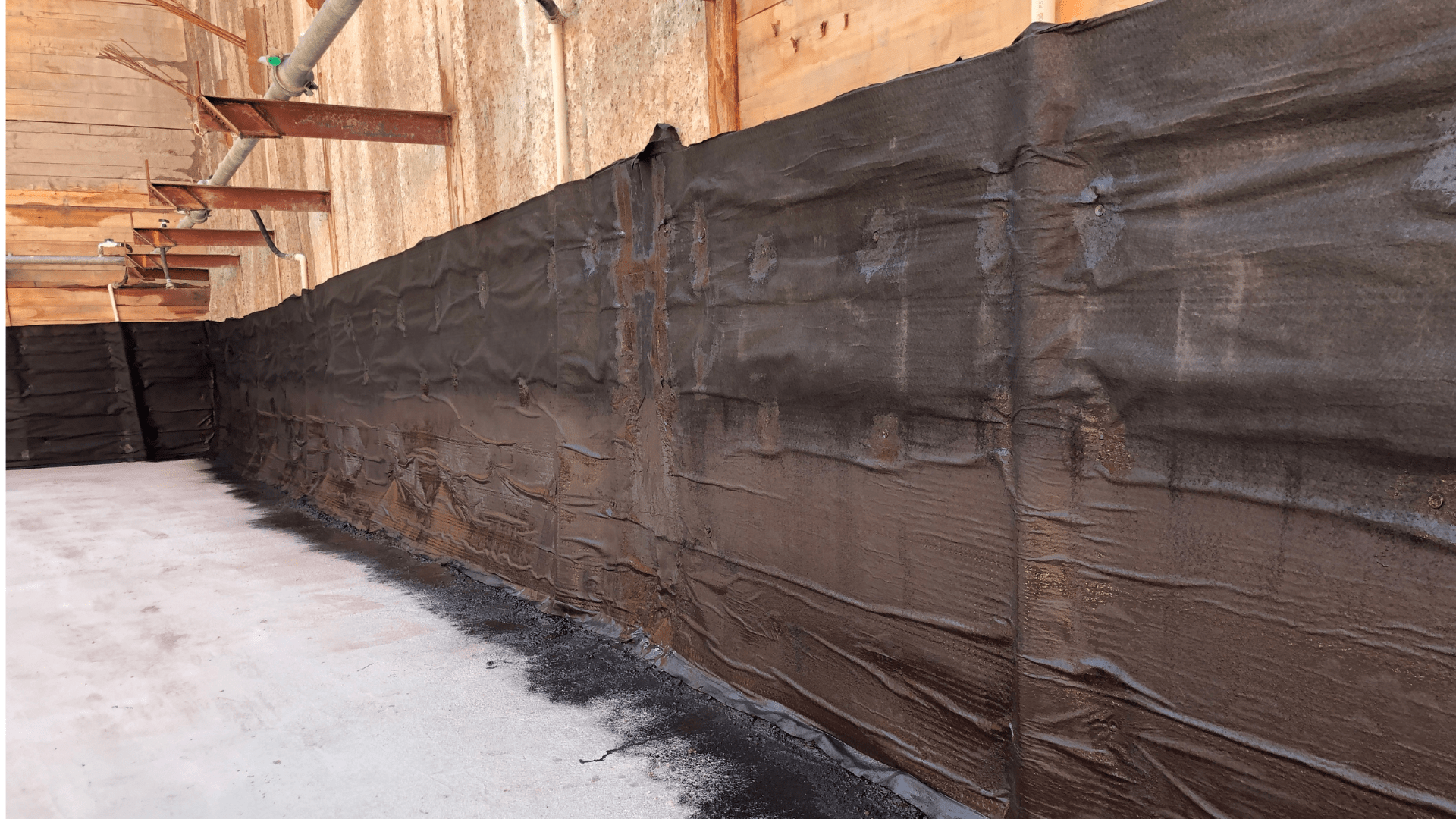 A construction site with a wall covered in black waterproofing or protective membrane. The wall runs alongside a concrete floor, with some debris and dust scattered at the base. Waterproofing Company | Basement Leak Repair | City Wide Group | Professional waterproofing company and basement leak repair in Toronto since 1961. | https://www.citywidegroup.com | +1 416-283-5500