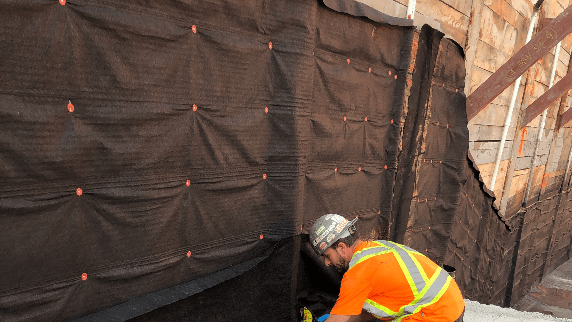 A construction worker in a safety vest and helmet installs black waterproofing material on a foundation wall at a building site, with wood forms and gravel visible around. Waterproofing Company | Basement Leak Repair | City Wide Group | Professional waterproofing company and basement leak repair in Toronto since 1961. | https://www.citywidegroup.com | +1 416-283-5500