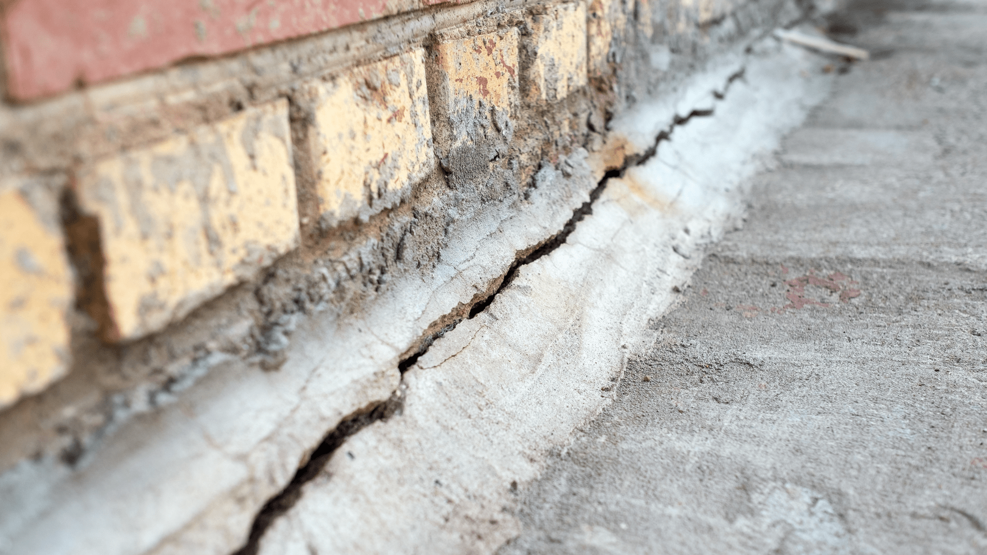 A close-up of a large horizontal crack at the base of a brick wall, where the concrete foundation meets the bricks. The crack runs along the length of the wall, indicating structural damage. Waterproofing Company | Basement Leak Repair | City Wide Group | Professional waterproofing company and basement leak repair in Toronto since 1961. | https://www.citywidegroup.com | +1 416-283-5500