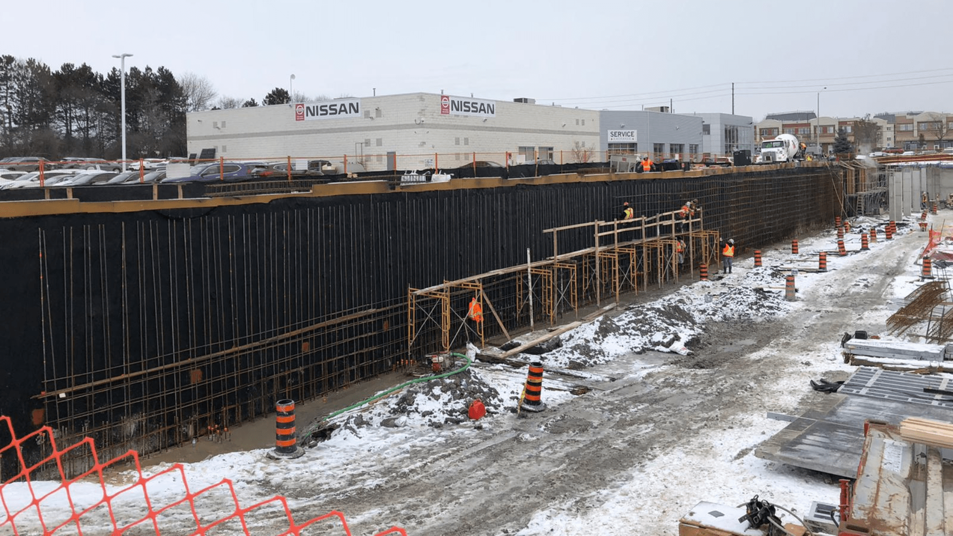 Construction workers in safety gear build and inspect a tall, black-coated concrete wall at a snowy construction site near a Nissan dealership. Scaffolding and orange traffic barrels are visible along the work area. Waterproofing Company | Basement Leak Repair | City Wide Group | Professional waterproofing company and basement leak repair in Toronto since 1961. | https://www.citywidegroup.com | +1 416-283-5500