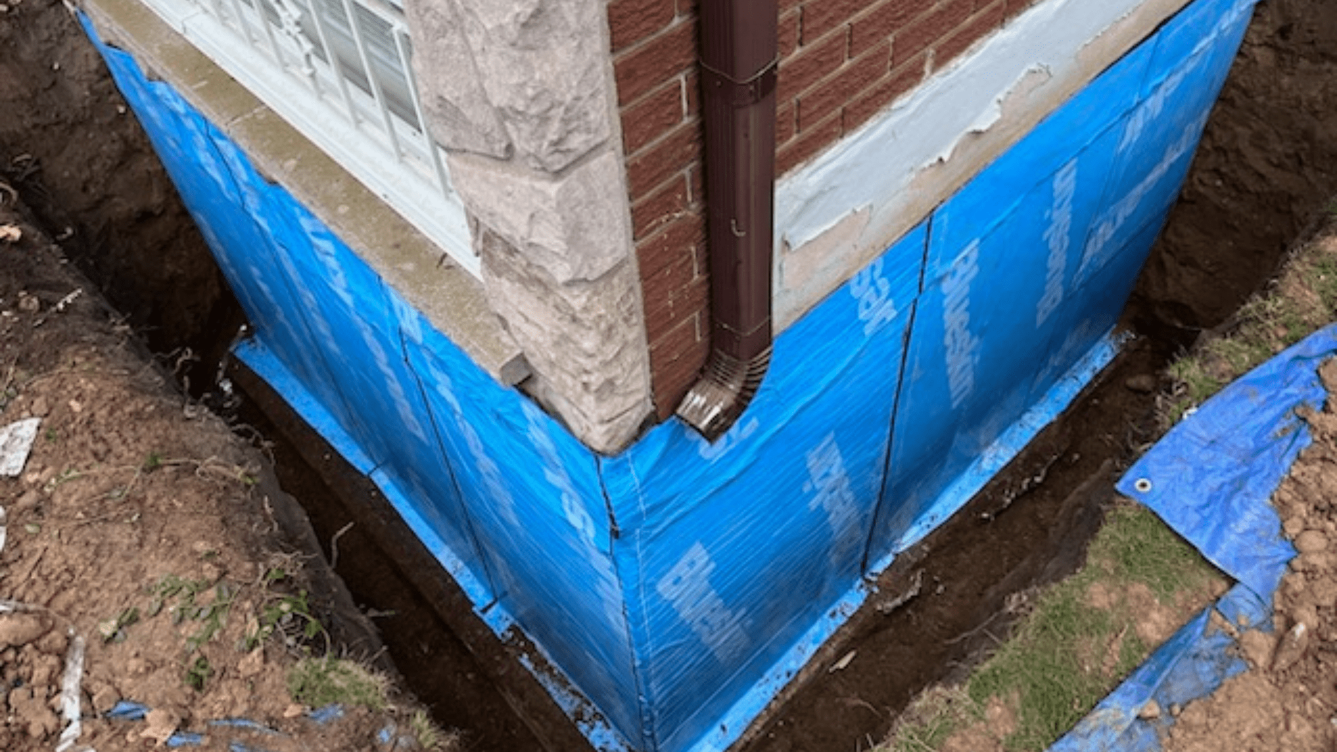 The corner of a house foundation is exposed, showing blue waterproofing membrane applied to the exterior walls, with a brown downspout attached. The surrounding soil has been dug out for what appears to be foundation repair. Waterproofing Company | Basement Leak Repair | City Wide Group | Professional waterproofing company and basement leak repair in Toronto since 1961. | https://www.citywidegroup.com | +1 416-283-5500