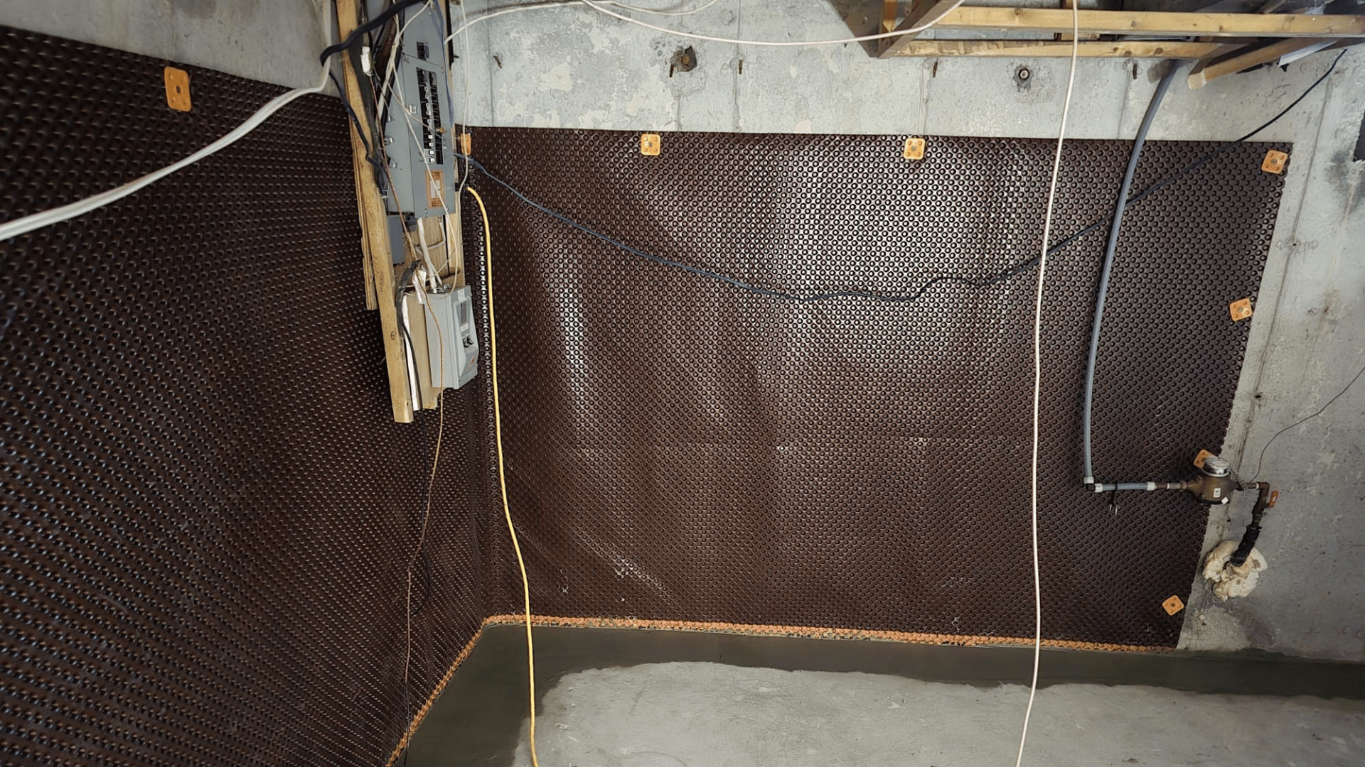 A basement corner with a brown dimpled waterproof membrane attached to the concrete walls, exposed wiring, utility piping, and unfinished flooring. Some wooden boards and electrical panels are mounted on the wall. Waterproofing Company | Basement Leak Repair | City Wide Group | Professional waterproofing company and basement leak repair in Toronto since 1961. | https://www.citywidegroup.com | +1 416-283-5500